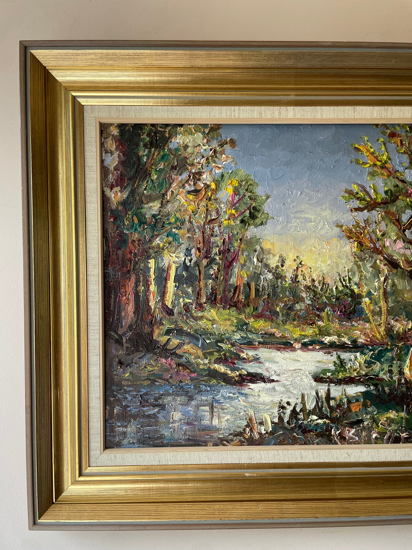 Impasto oil painting of a sunlit woodland stream framed by trees under a pale blue sky, in a gold gilt frame