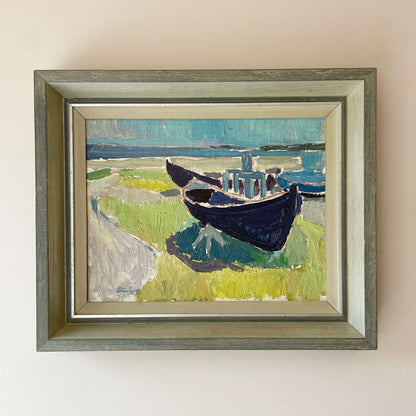 Mid-century Swedish oil painting of two blue fishing boats beached on green grass beside a turquoise sea, in a silvered grey-green wooden frame.