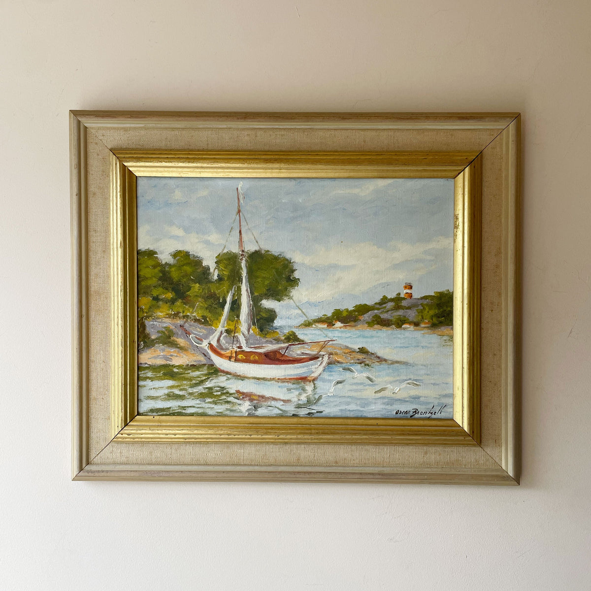 Oil painting of a white sailing boat moored by a rocky wooded shore, with a red and white lighthouse and gulls, in a linen and gilt frame