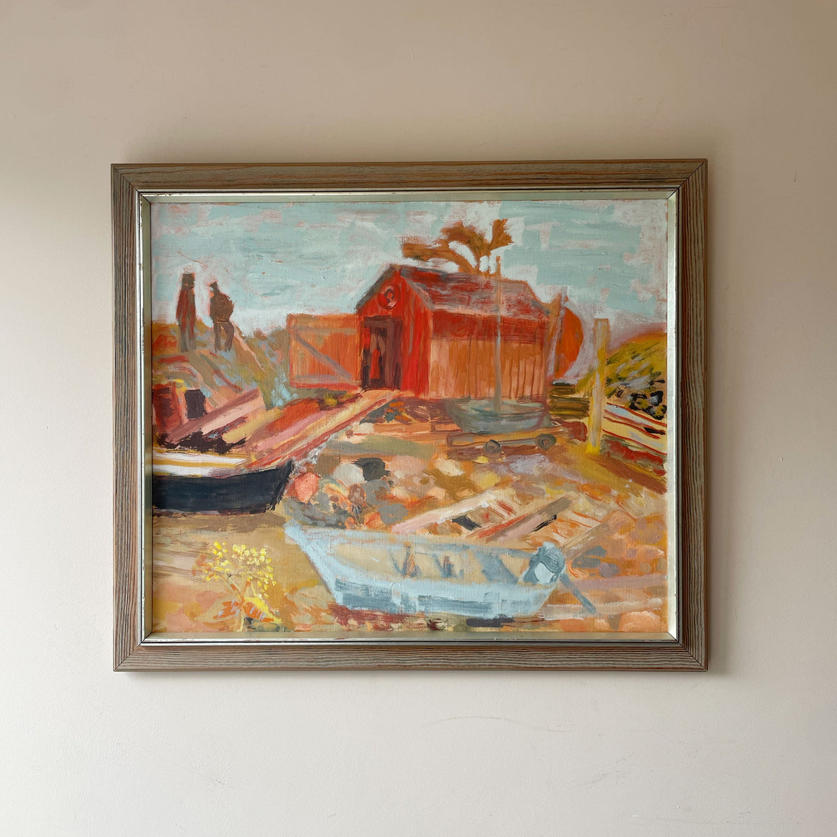 Mid century oil painting of boatyard  on a rocky shore, with two figures and a pale blue sky, in a limed wood frame. Red and orange.